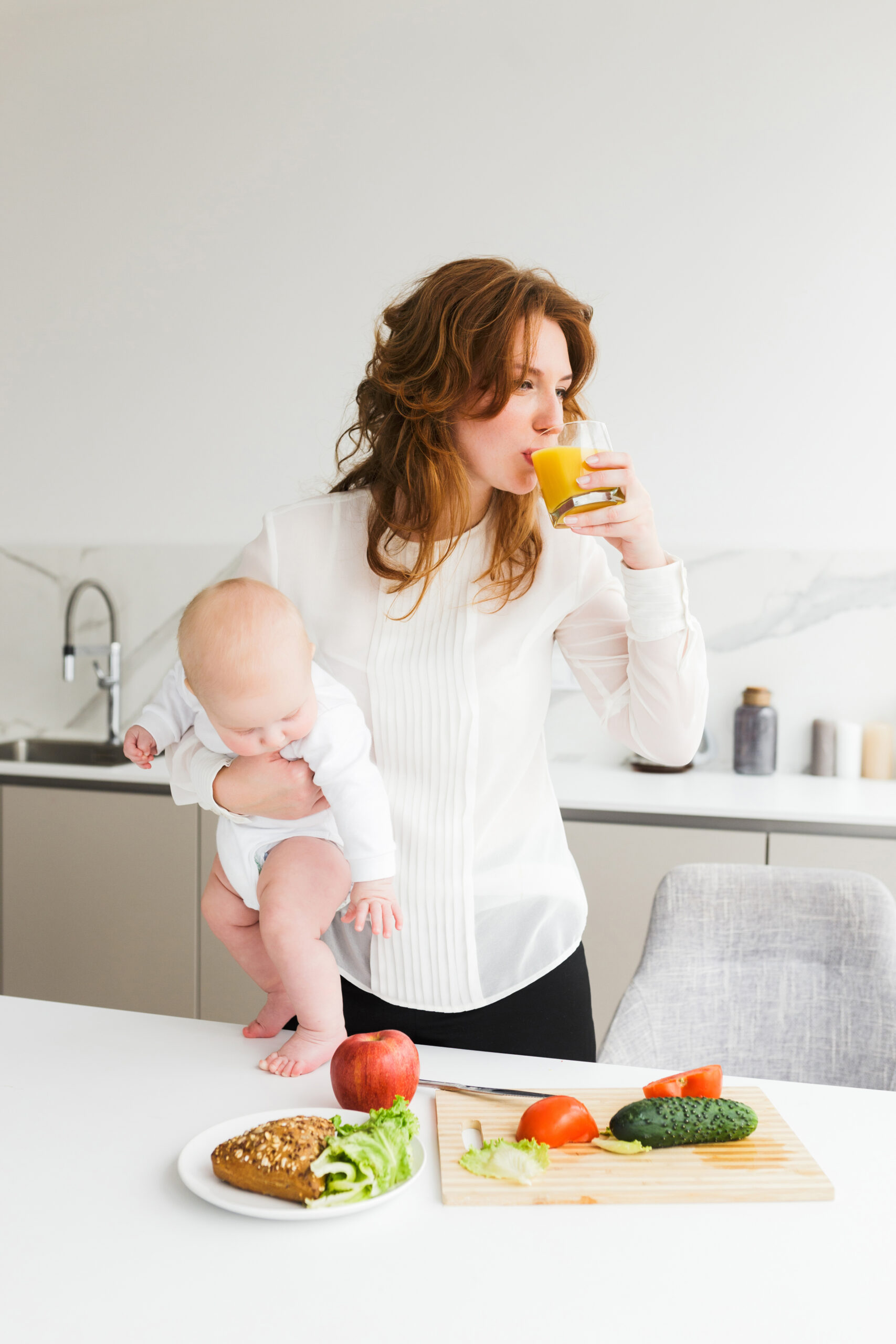 Young pretty woman standing and holding her little baby while drinking juice and cooking on kitchen